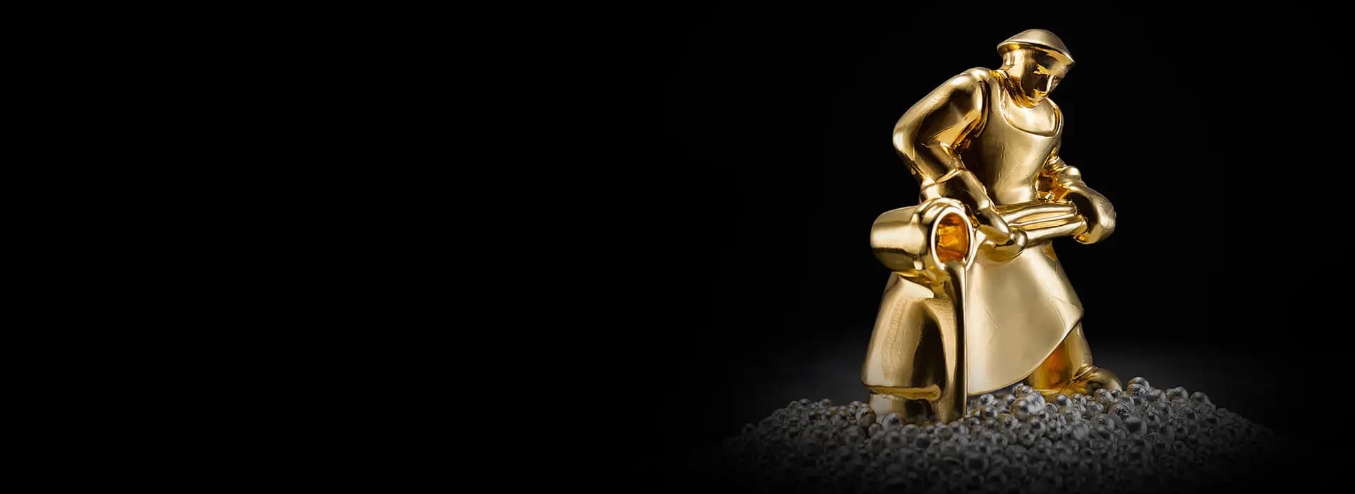 Golden statue of a person in an apron pouring molten metal, surrounded by small pellets, set against a black background.